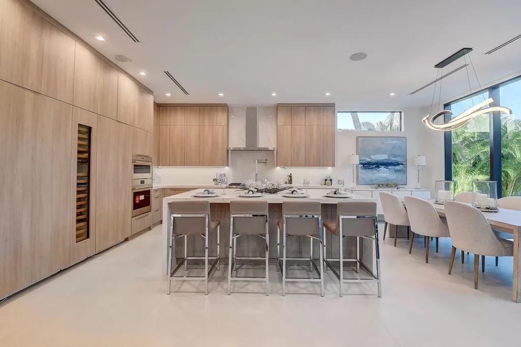 Modern kitchen and dining area with sleek wooden cabinetry, an island with bar stools, and an adjoining dining table set under pendant lighting.