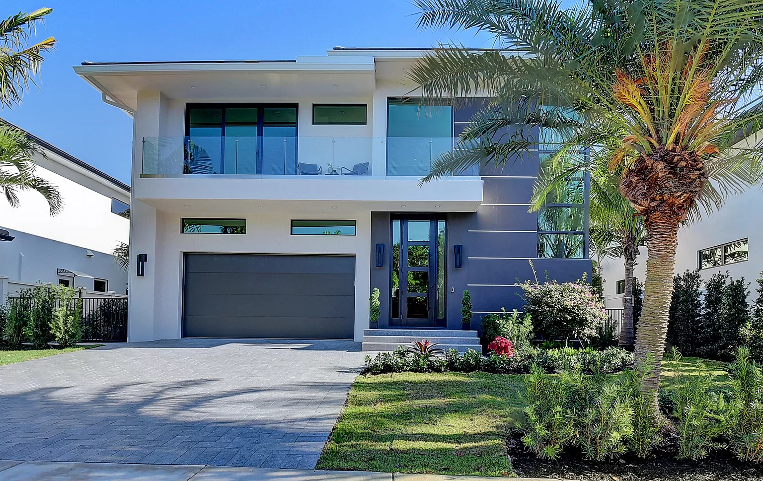 Modern two-story home with clean lines, featuring a large glass balcony, a two-car garage, and landscaped with palm trees.
