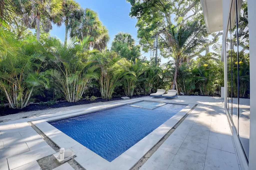 Modern backyard with a rectangular swimming pool surrounded by lush greenery and adjacent to a house with large glass doors.