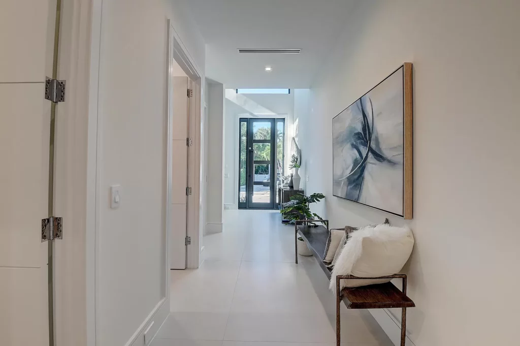 A bright hallway with modern decor featuring a large artwork on the wall, a wooden bench with a white throw, and a potted plant near an open door at the end.