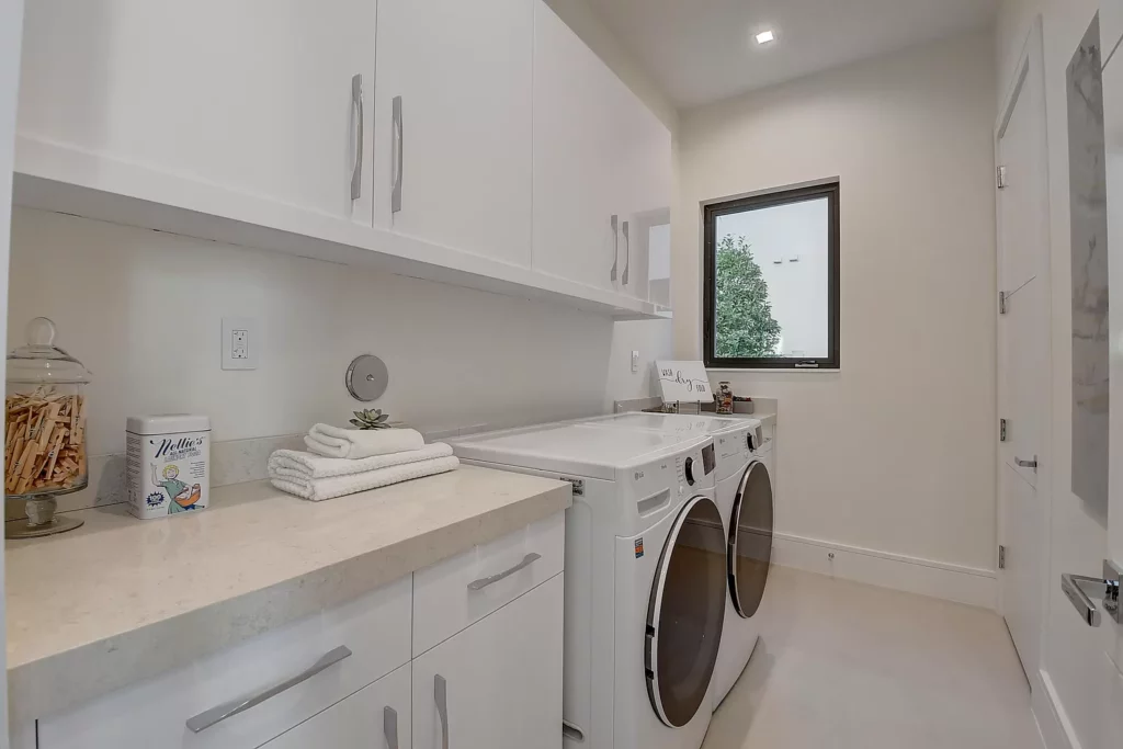 A modern laundry room with white appliances, cabinets, and a window overlooking greenery.