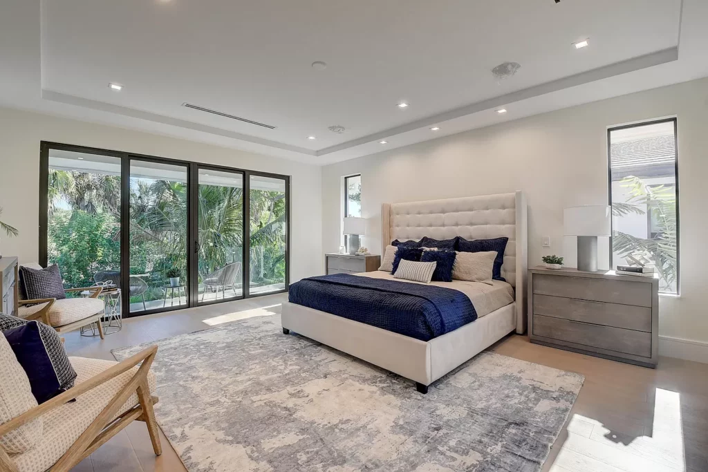 A modern bedroom with a large upholstered bed, sliding glass doors, and a tropical view.